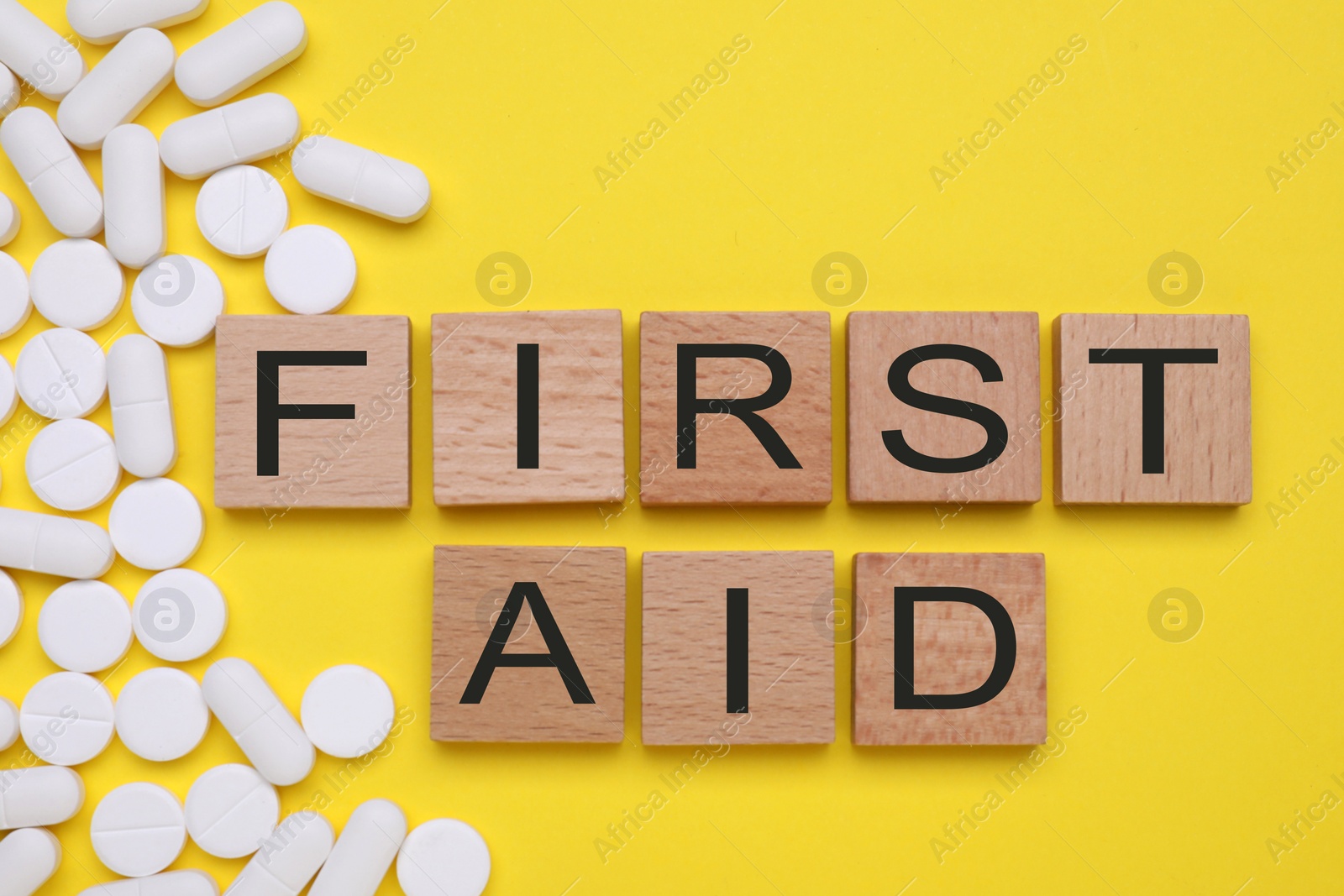 Photo of Words First Aid made of wooden cubes and pills on yellow background, flat lay