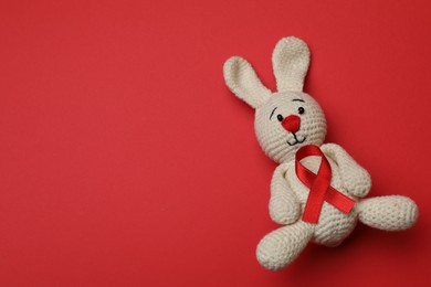 Cute knitted toy bunny with ribbon on red background, top view and space for text. AIDS disease awareness