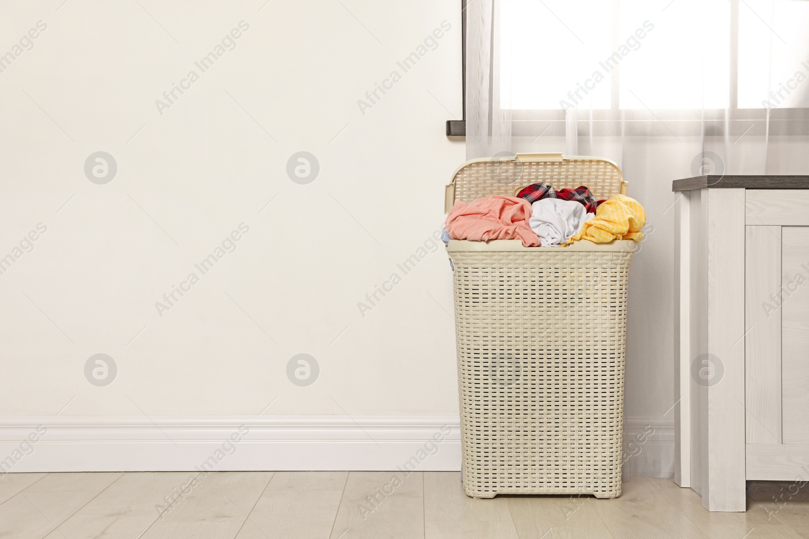 Photo of Plastic laundry basket full of dirty clothes on floor near light wall in room. Space for text