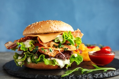 Tasty burger with bacon on slate plate against color background