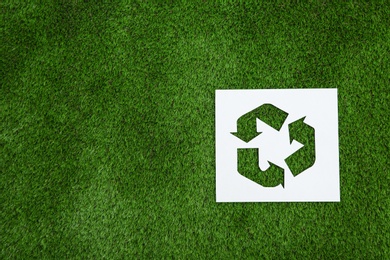 Sheet of paper with cutout recycling symbol on green grass, top view. Space for text