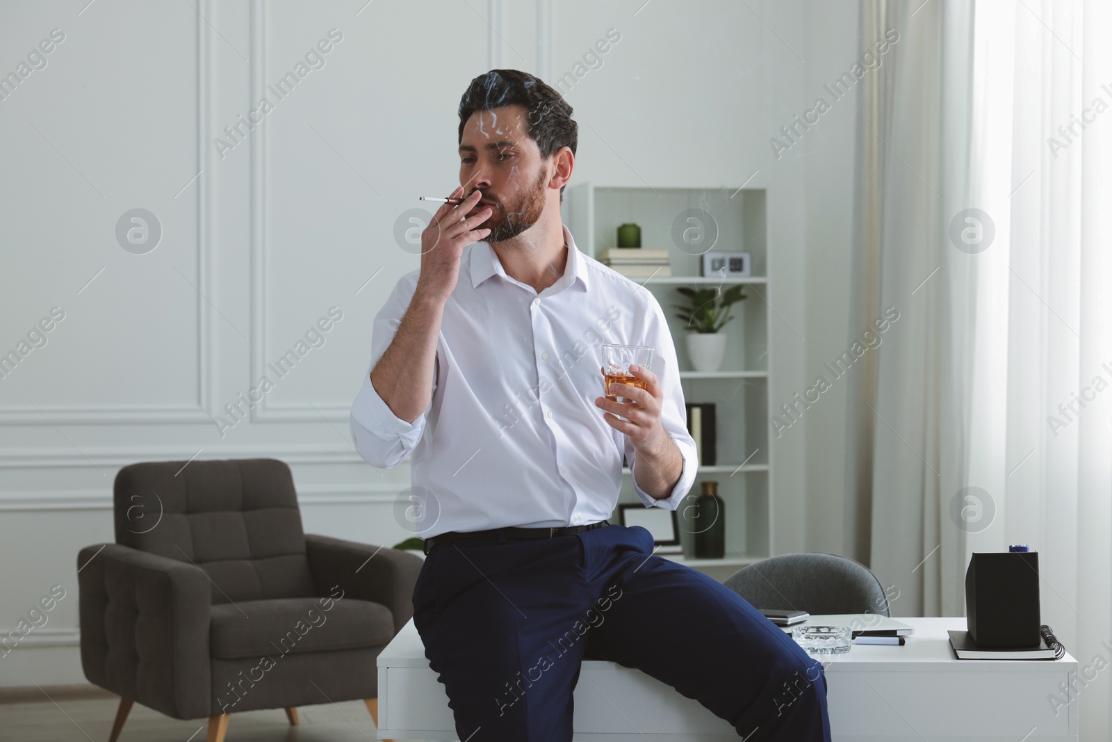 Photo of Man using cigarette holder for smoking and holding glass of whiskey in office