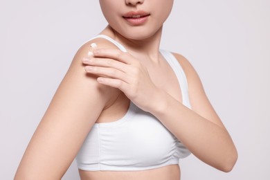 Photo of Young woman applying body cream onto shoulder on light grey background, closeup