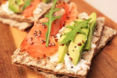 Photo of Fresh crunchy crispbreads with cream cheese, salmon, avocado and arugula on wooden board, closeup