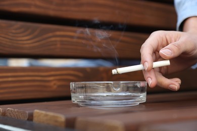 Photo of Woman holding cigarette over glass ashtray on bench outdoors, closeup
