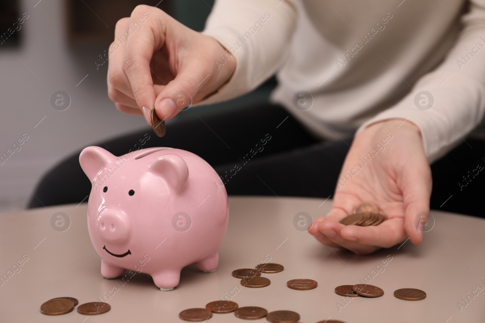 Photo of Woman putting coin into piggy bank at table indoors, closeup