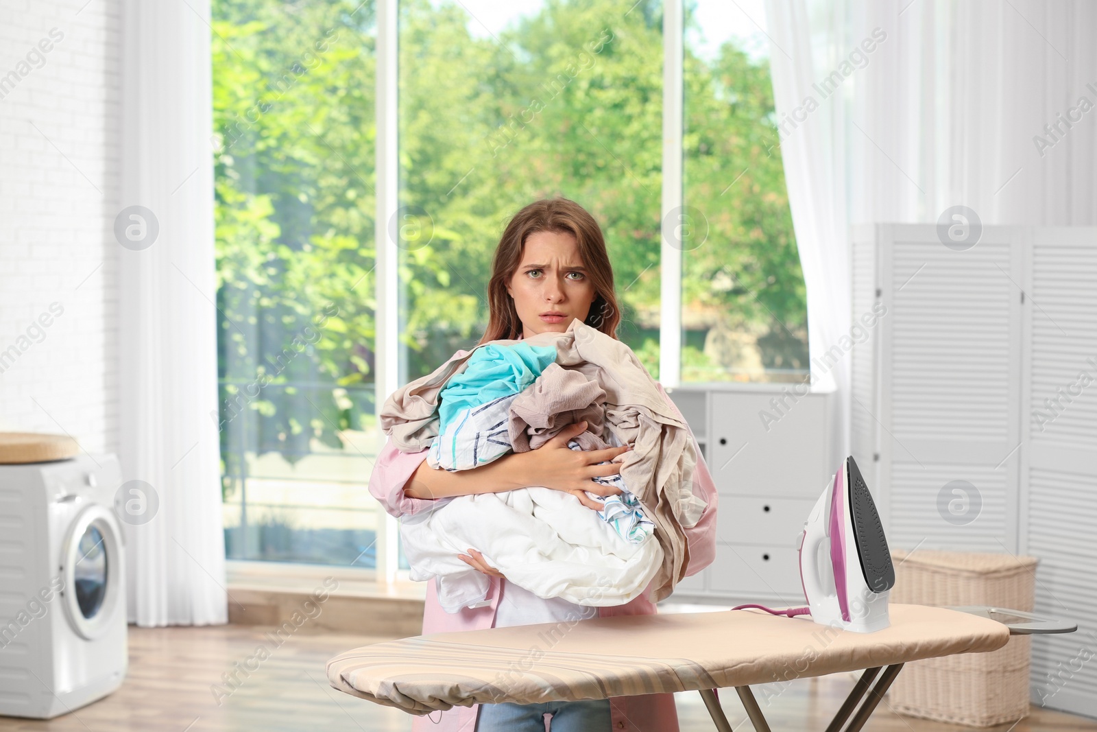 Photo of Tired girl holding unfolded laundry near ironing board at home