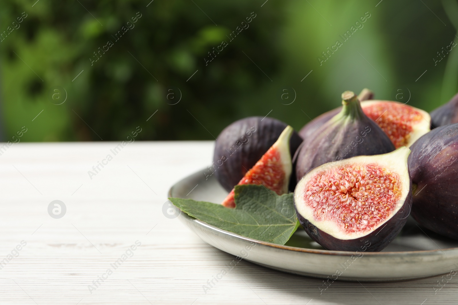 Photo of Whole and cut ripe figs with leaf on white wooden table against blurred green background, closeup. Space for text