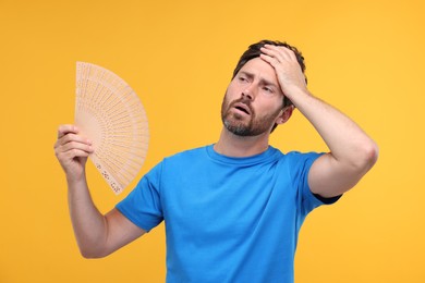 Unhappy man with hand fan suffering from heat on orange background