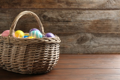 Photo of Wicker basket with painted Easter eggs on table against wooden background, space for text