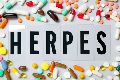 Photo of Word Herpes surrounded by different pills on white background, top view