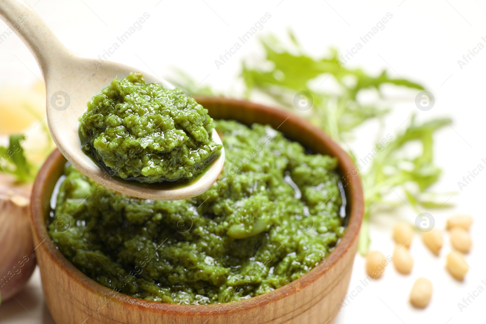 Photo of Spoon of tasty arugula pesto near bowl with sauce, closeup