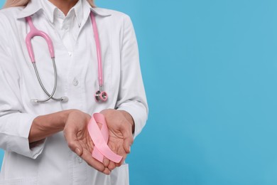 Doctor with stethoscope holding pink ribbon on light blue background, closeup and space for text. Breast cancer awareness