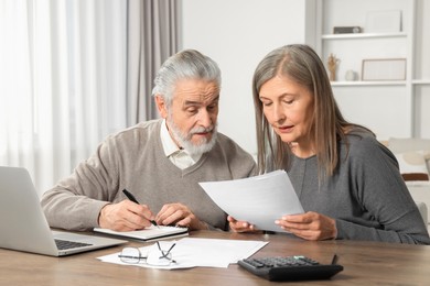 Photo of Elderly couple with papers and laptop discussing pension plan at wooden table in room