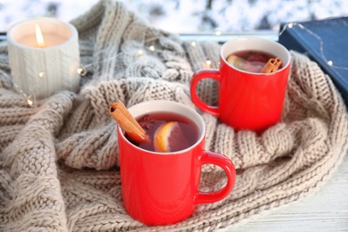 Photo of Cups of hot winter drink with scarf on window sill indoors