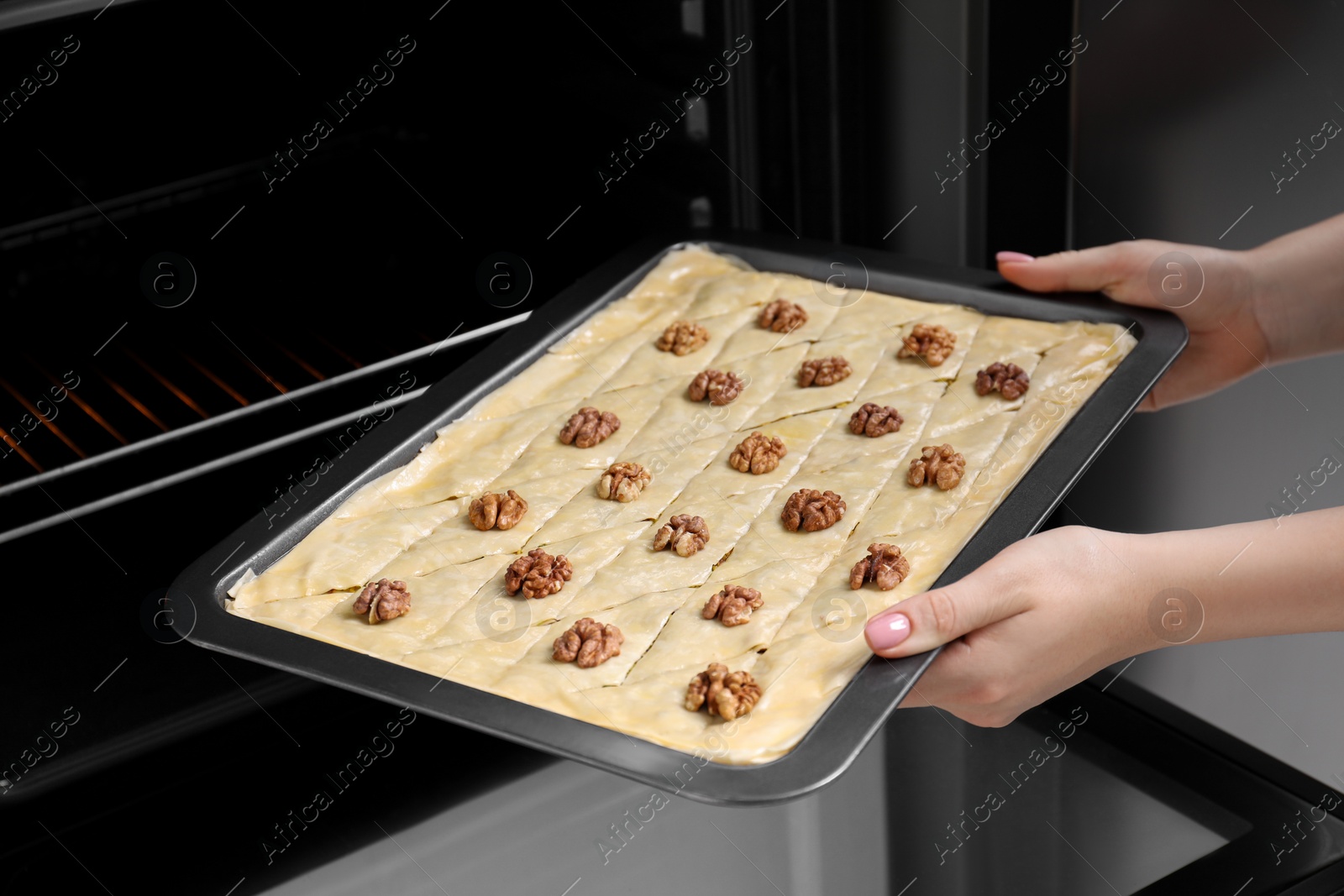 Photo of Woman putting baking pan with baklava into oven, closeup