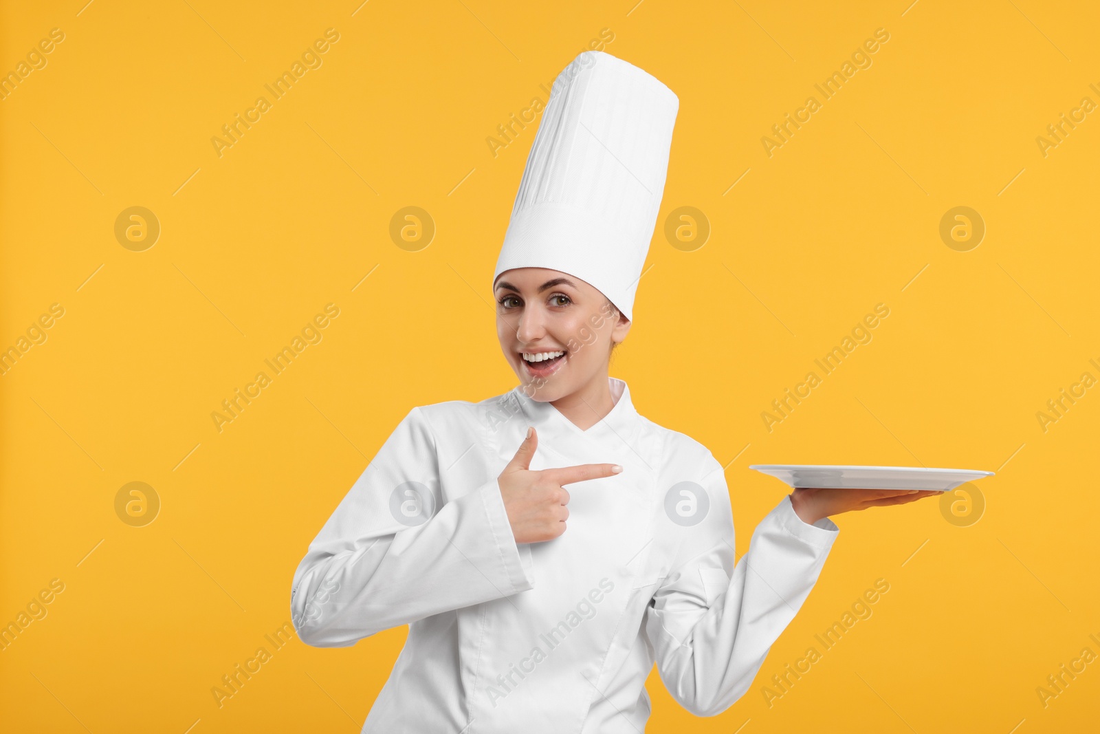 Photo of Happy professional confectioner in uniform pointing at empty plate on yellow background