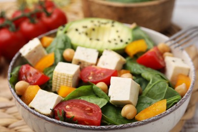 Photo of Bowl of tasty salad with tofu and vegetables on wicker mat, closeup