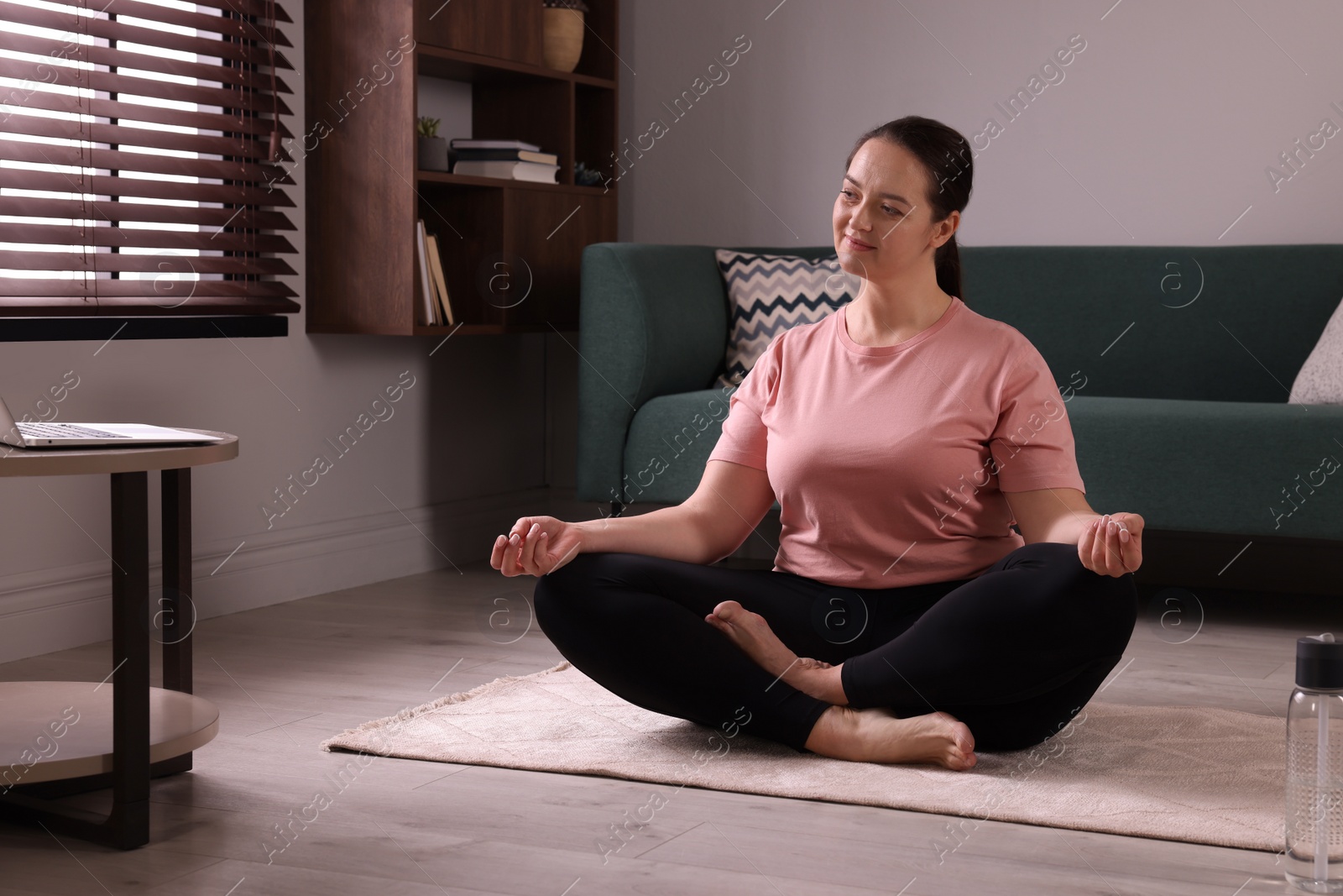 Photo of Overweight woman meditating on rug while watching online class at home
