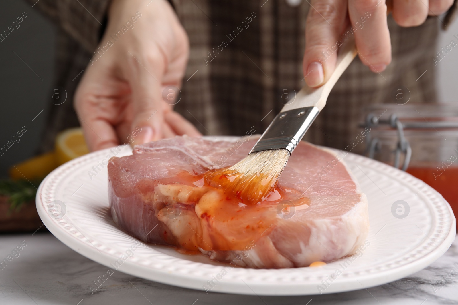 Photo of Woman spreading marinade onto raw meat with basting brush at white marble table, closeup