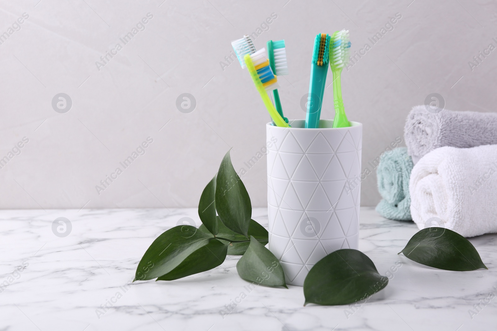 Photo of Colorful plastic toothbrushes in container and green leaves on white marble table, space for text