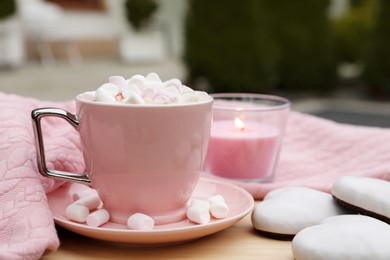 Photo of Cup of tasty cocoa with marshmallows, pink sweater, cookies and burning candle on wooden table outdoors