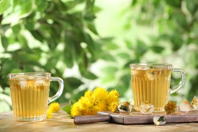 Photo of Delicious fresh tea, dandelion flowers and ice cubes on wooden table against blurred background