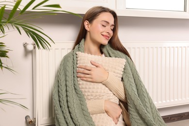 Photo of Woman with blanket hugging pillow near heating radiator indoors