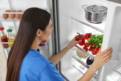 Photo of Young woman taking red bell pepper out of refrigerator