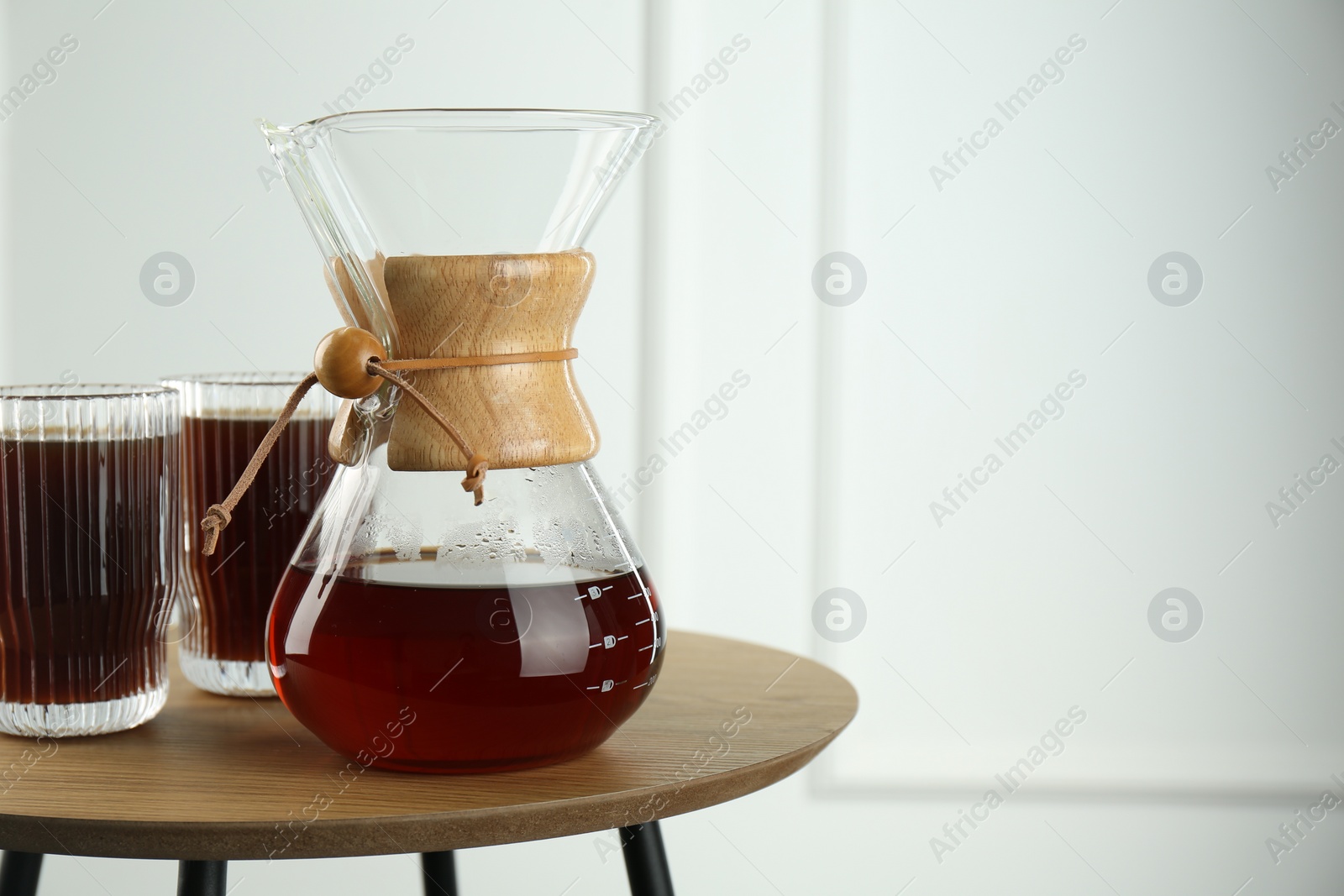 Photo of Glass chemex coffeemaker and glasses of coffee on wooden table against white wall, space for text