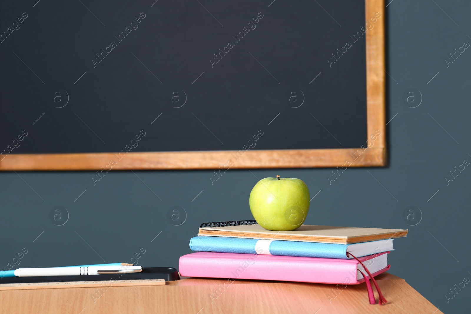 Photo of Wooden school desk with stationery and apple near blackboard on grey wall