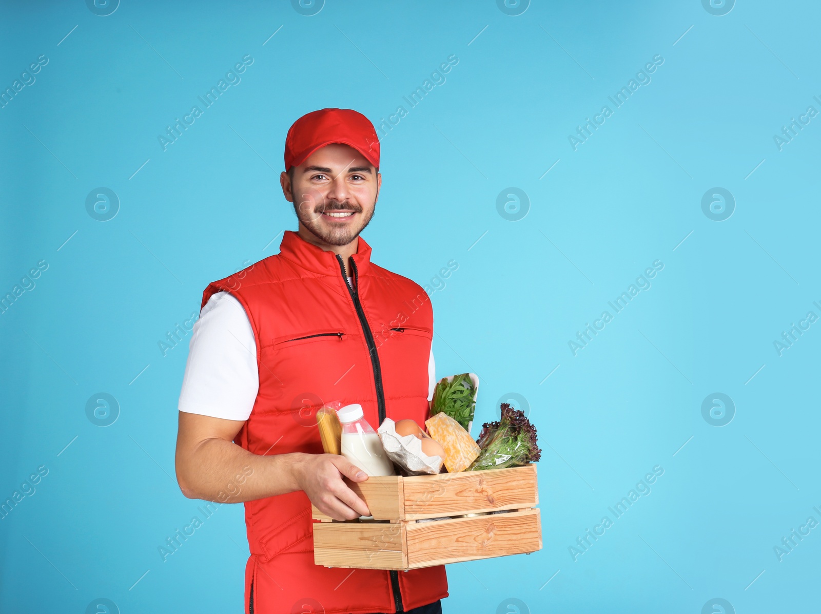 Photo of Food delivery courier holding wooden crate with products on color background. Space for text