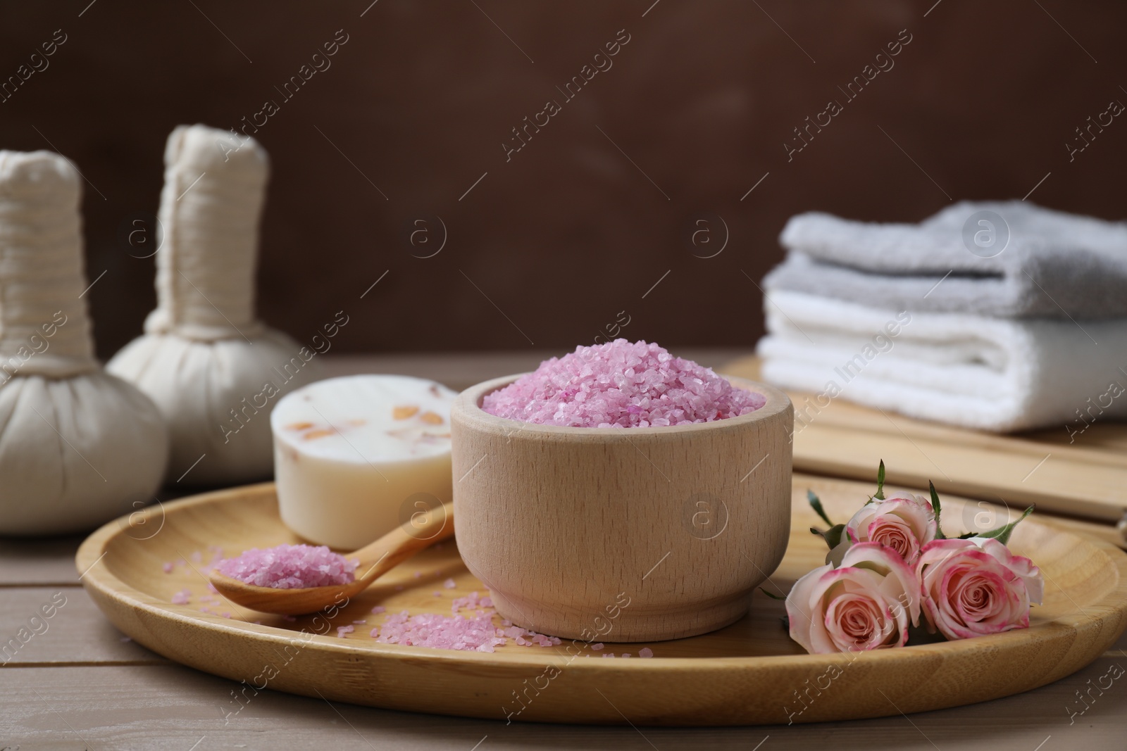 Photo of Bowl of pink sea salt, roses and herbal massage bags on wooden table