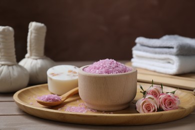 Bowl of pink sea salt, roses and herbal massage bags on wooden table