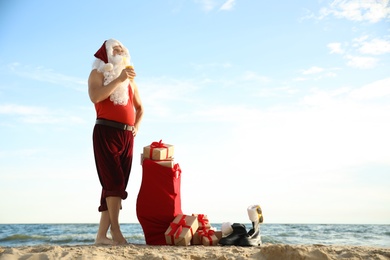 Photo of Santa Claus with cocktail and bag of presents on beach, space for text. Christmas vacation