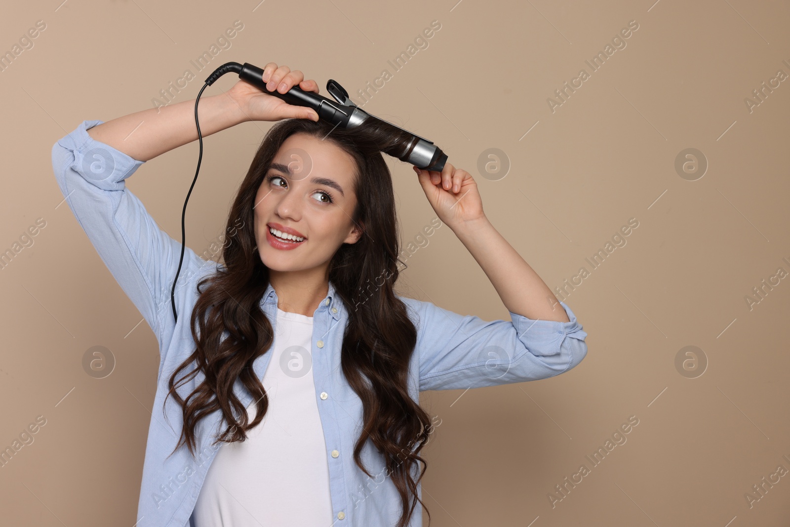 Photo of Happy woman using curling hair iron on beige background. Space for text