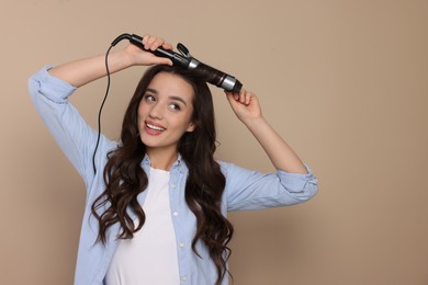 Happy woman using curling hair iron on beige background. Space for text