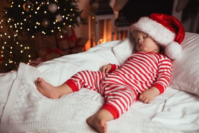 Photo of Baby in Christmas pajamas and Santa hat sleeping on bed indoors