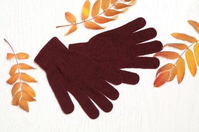 Stylish woolen gloves and dry leaves on white wooden table, flat lay