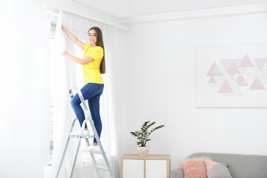 Young woman hanging window curtain in room. Space for text