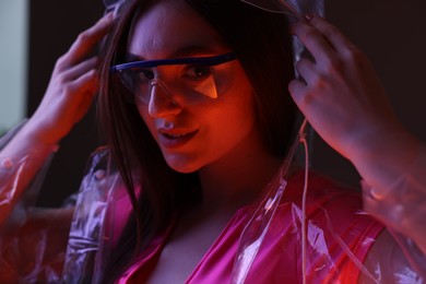 Fashionable portrait of beautiful woman wearing transparent coat and glasses on dark background in neon lights, closeup