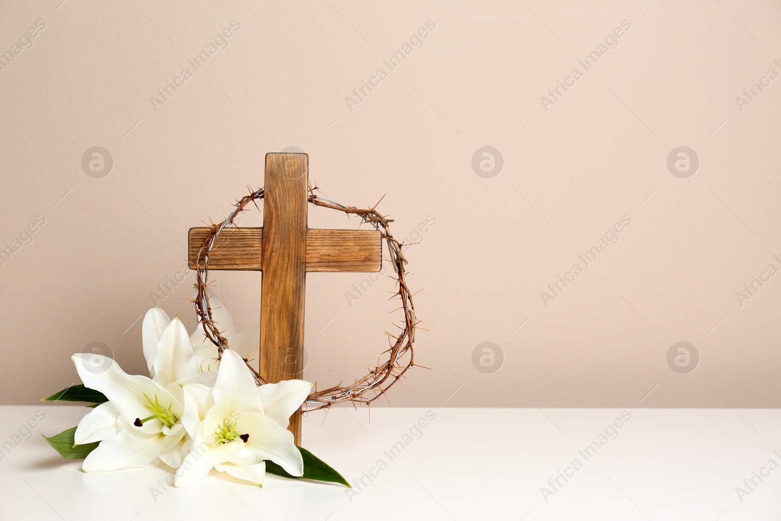 Photo of Wooden cross, crown of thorns and blossom lilies on table against color background, space for text