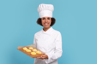 Happy female chef in uniform holding board with cookies on light blue background