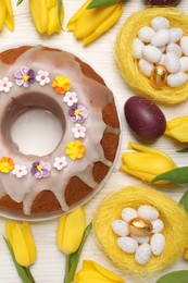 Flat lay composition of delicious Easter cake decorated with sprinkles, eggs and beautiful tulips on white wooden table