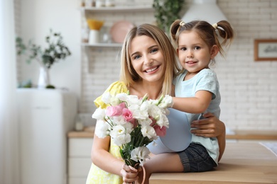 Photo of Young mother with bouquet and gift box hugging her little daughter in kitchen