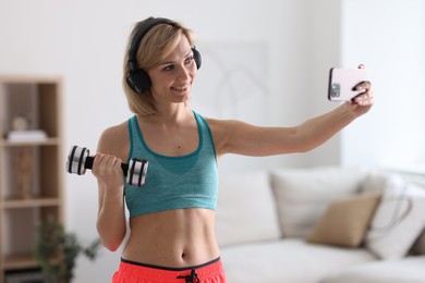 Photo of Smiling sports blogger holding dumbbell while streaming online fitness lesson with smartphone at home