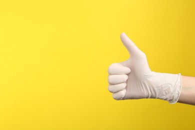 Photo of Person in medical gloves showing thumb up on yellow background, closeup of hand. Space for text