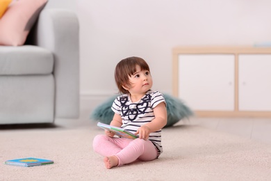 Photo of Cute baby girl with book sitting on floor at home