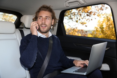 Photo of Young handsome man talking on phone while using laptop in car