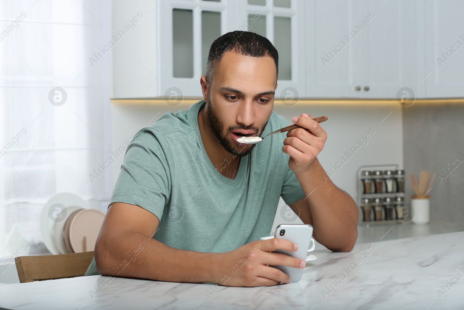 Photo of Young man using smartphone while having breakfast at white marble table in kitchen. Internet addiction
